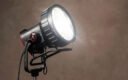 Увімкнений моноблок Aputure STORM 700x з лінзою Френеля CF10 Fresnel на сталевій стійці, розгорнутий під кутом на бежево-коричневому фоні: металева ліра з фіксатором нахилу та кабель до виносного блока керування