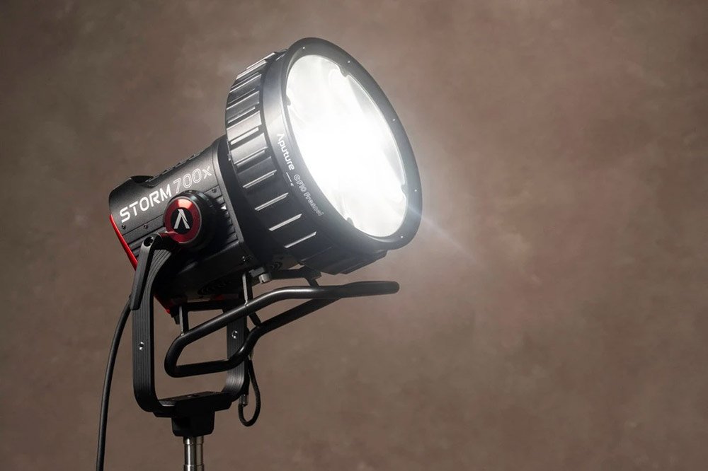 Увімкнений моноблок Aputure STORM 700x з лінзою Френеля CF10 Fresnel на сталевій стійці, розгорнутий під кутом на бежево-коричневому фоні: металева ліра з фіксатором нахилу та кабель до виносного блока керування